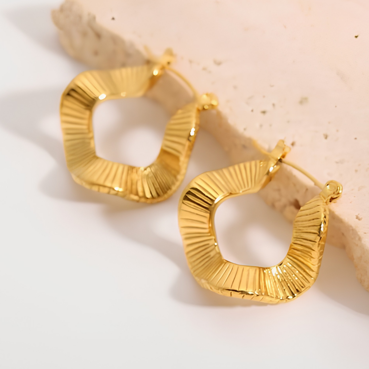 A pair of textured gold hoop earrings with a sunburst or ribbed pattern, displayed on a light beige stone slab against a clean white background.