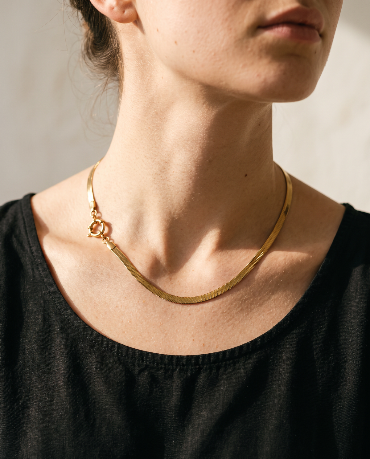 A close-up of a model wearing a thin gold snake chain necklace with a small interlocking double-heart charm, paired with a black top against a neutral background.