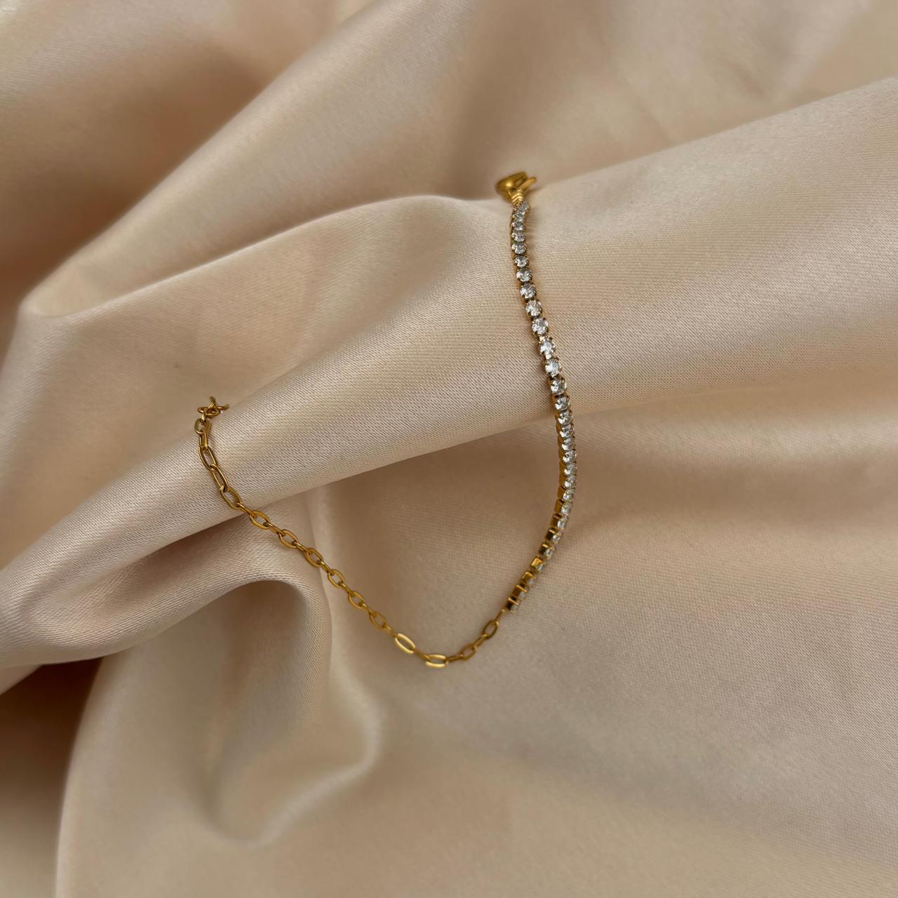 Gold bracelet with half clear stones and half paperclip chain on a beige fabric background