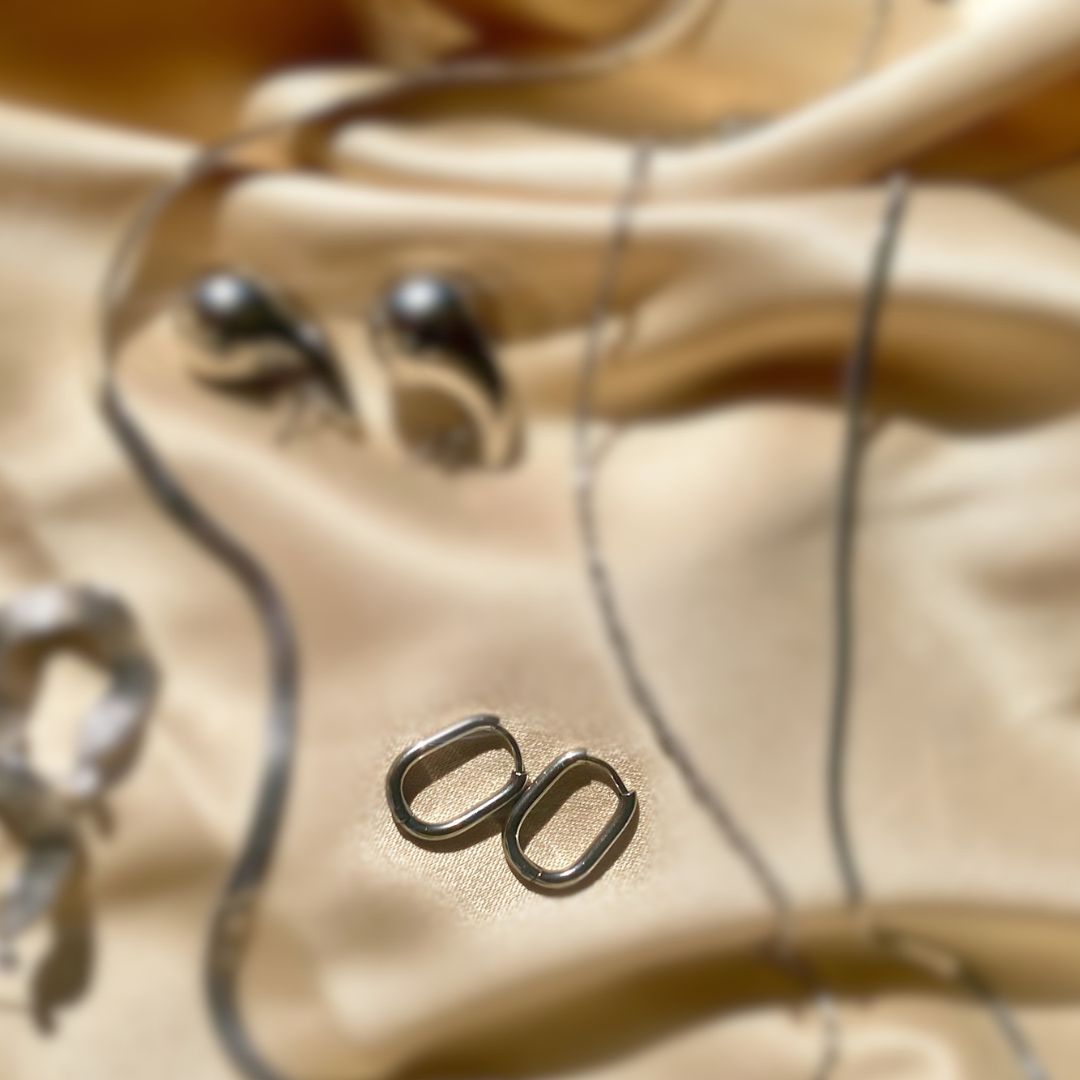 A close-up of a pair of silver oval hoop earrings resting on draped beige satin fabric.