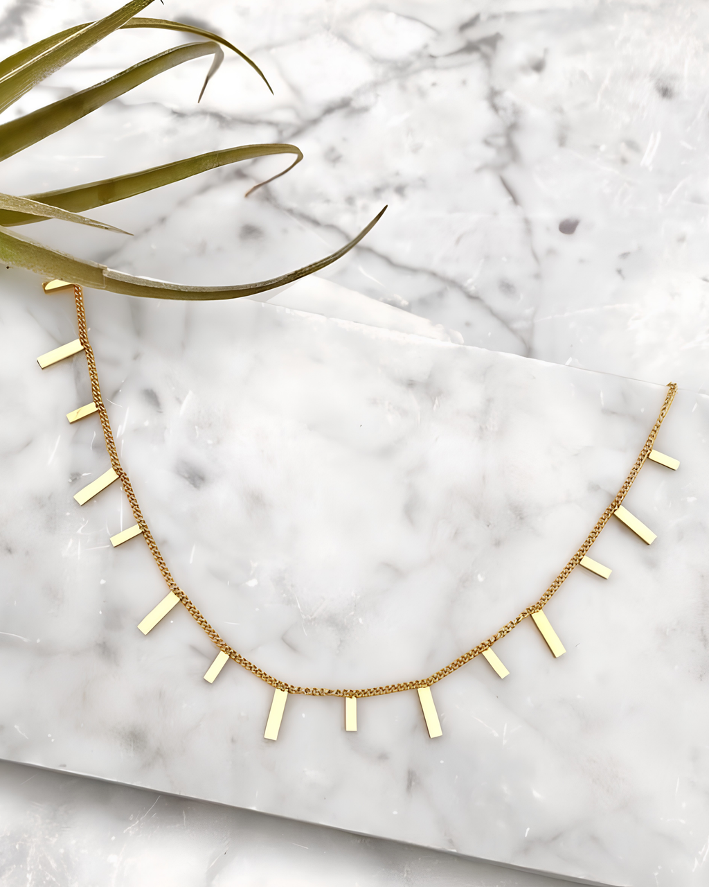 A delicate gold chain necklace featuring small, evenly spaced rectangular bar charms, displayed on a white marble slab with a green plant leaf in the corner.