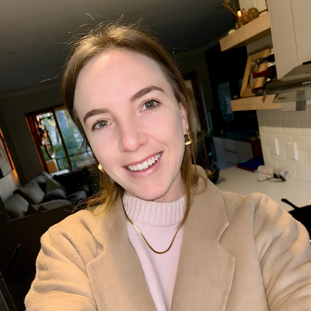 A smiling woman taking a selfie in a home kitchen, wearing a beige fuzzy coat over a light pink turtleneck sweater.
